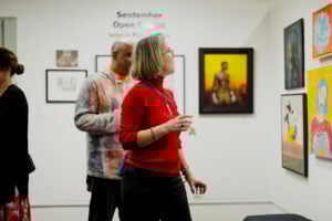 A woman in a red shirt and a man in a colorful sweater look at brightly colored paintings on the wall.